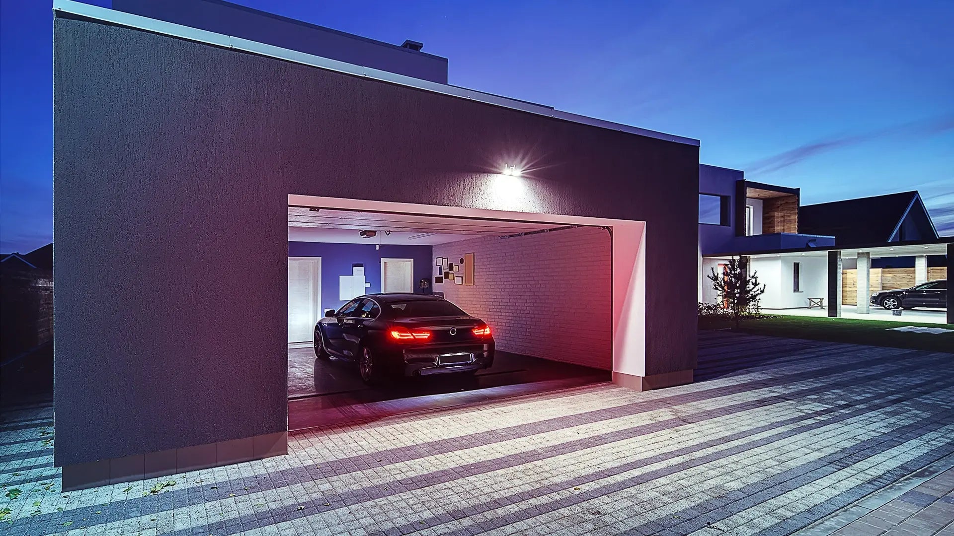 Modern home at night with an open garage door and a car backing in, lit by a smart exterior light.