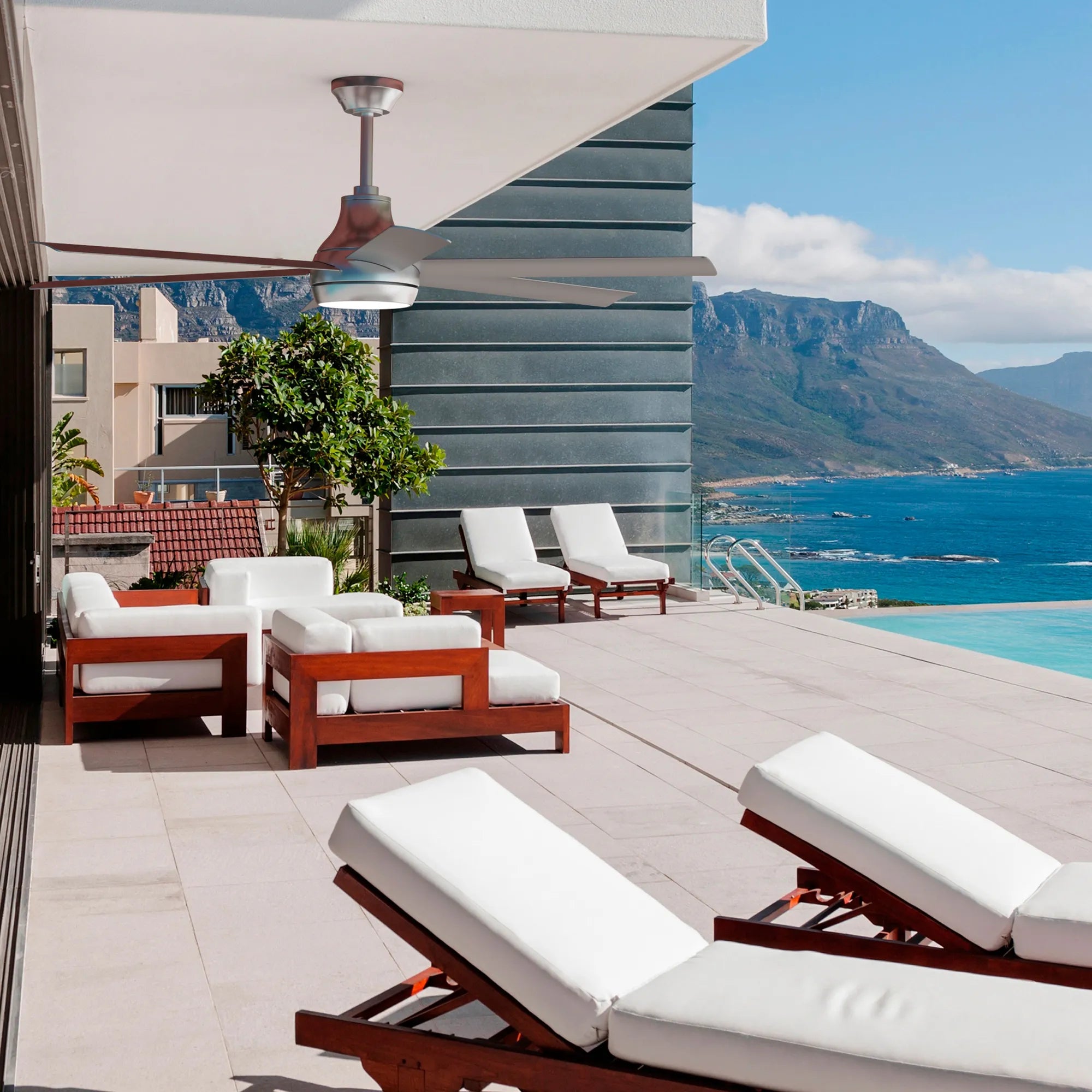 Silver Bresio Smart Outdoor Ceiling Fan mounted above a luxury patio with white cushioned lounge chairs and oceanfront view.