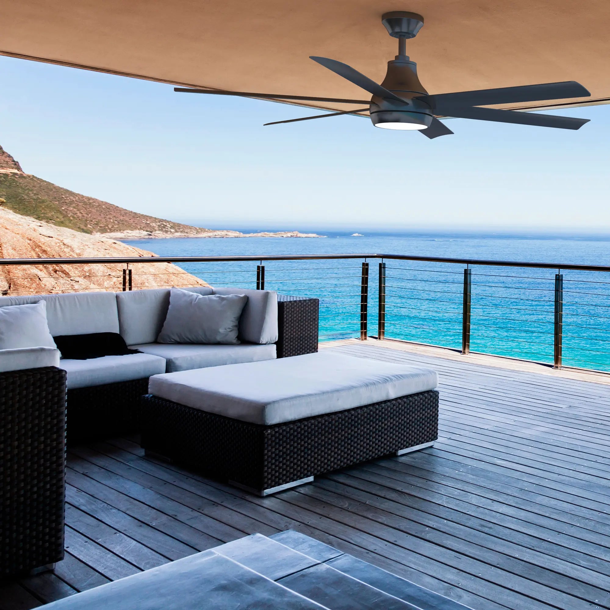 Outdoor seating area with lounge chairs under a covered deck featuring the Bresio Smart Ceiling Fan in pearl black finish, overlooking the ocean.