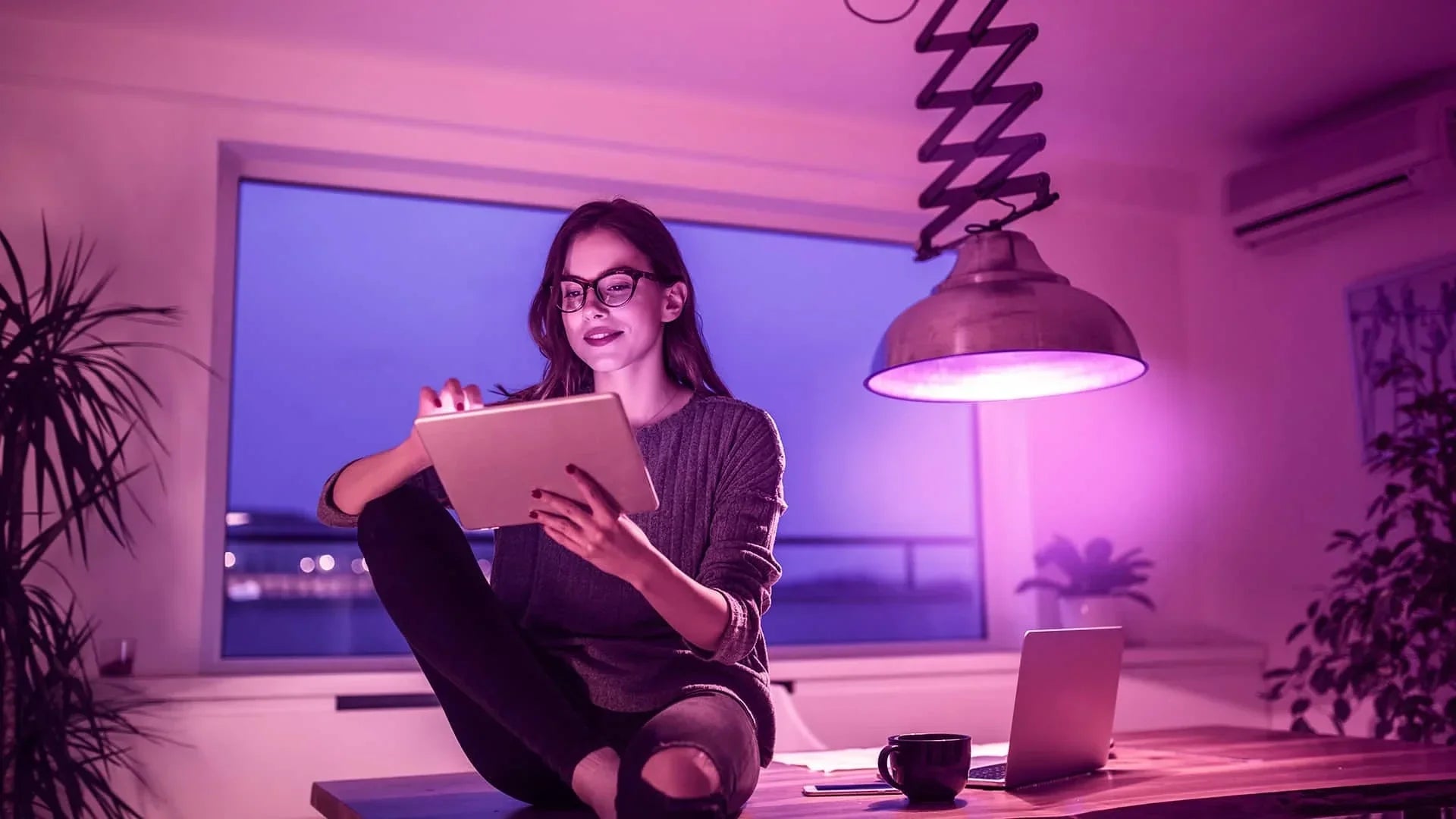 Woman sitting on a desk using a tablet under a purple-tinted smart light, with a laptop and coffee nearby, illustrating the mood-enhancing and productivity benefits of smart lighting.